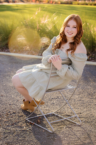 Graceful high school senior in sage green dress sitting in chair with sunflare and layered textures of grass and light