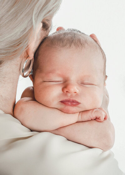 A newborn baby resting his chin on hes arms while resting on mom's shoulder, while mom is looking towards the white backdrop.