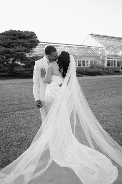 Timeless black and white portrait of a bride and groom embracing in front of a Paris glasshouse, captured by luxury destination wedding photographer Madison