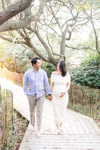 sun-drenched portrait of an expecting mom and husband walking hand in hand under shady oak tress in virginia beach during maternity photos