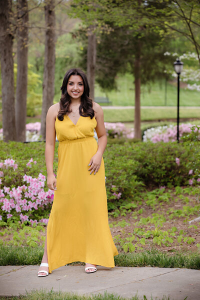 Graceful high school senior in sage green dress sitting in chair with sunflare and layered textures of grass and light