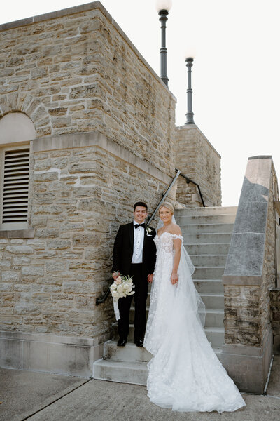 Bride and groom pose by stone staircase in elegant wedding attire.