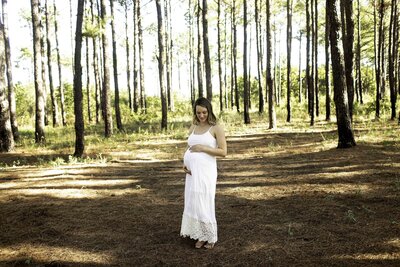 Expecting mother posing gracefully in a serene pine tree forest, glowing in natural light. Surrounded by tall, lush green trees, she gently cradles her baby bump, wearing a flowing dress that blends with the tranquil, earthy backdrop