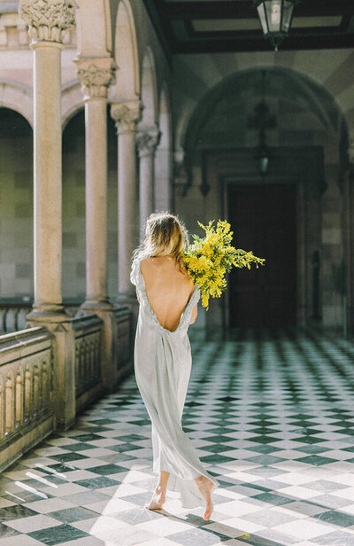 Femme de dos, pieds nus, portant une longue robe fluide blanche, bouquet de mimosas sur l'épaule, marchant sur un carrelage à carreaux dans un corridor entouré de colonnes.