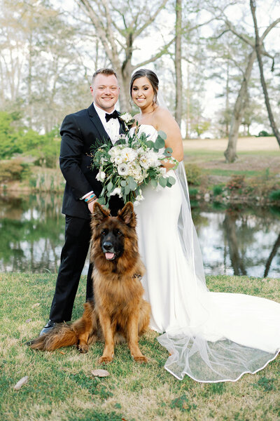 man in black tux next to woman in white dress with flowers with a large dog sitting in front of them