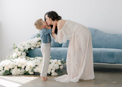 woman wearing a beautiful white flowy dress bending down and kissing her young son in front of a blue couch with a white floral arrangement in front of the couch- Maegan R Photography