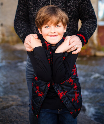 A smiling boy in a red-patterned vest hugged by his father, standing near water at Babcock Mill.