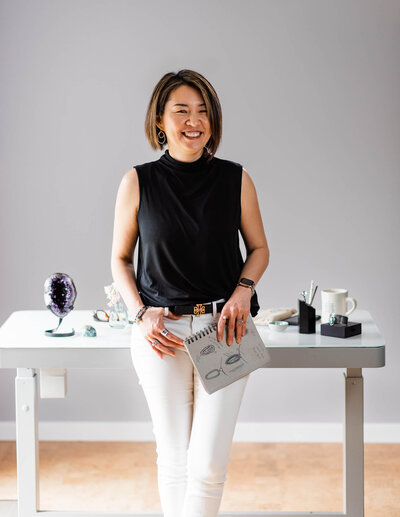 Female jeweler stands in front of her desk holding her sketchbook