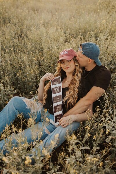 A couple is sitting in a field, with 'Mom' and 'Dad' hats on, holding an ultrasound, for their pregnancy reveal photoshoot
