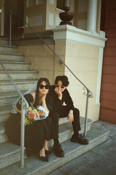 A couple sits on a stoop in San Francisco, they are dressed in all black with sunglasses and a floral bouquet for their engagement photos