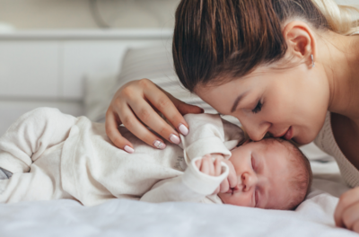 A newborn girl sleeps peacefully in a white bed, dressed in a cute pink pajama, as her mom gently kisses her face, capturing a tender moment at a Texas postnatal retreat.