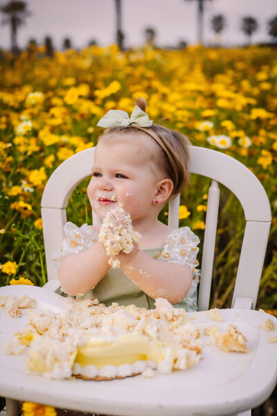 Cake Smash Photographer, a baby girl sits on a flowery chair and holds a wooden carving that reads "one"