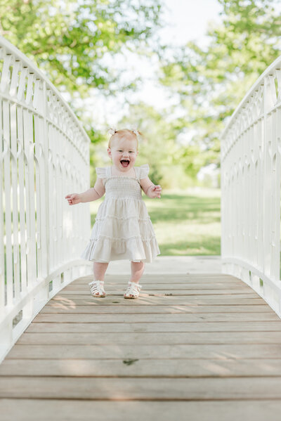 Smiling toddler girl walking across a white wooden bridge in a white dress during a bright and joyful Kansas City family photography session.