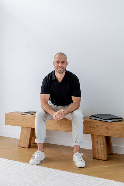 Jon Moses wearing a blue polo shirt, beige pants sitting on a wooden stool