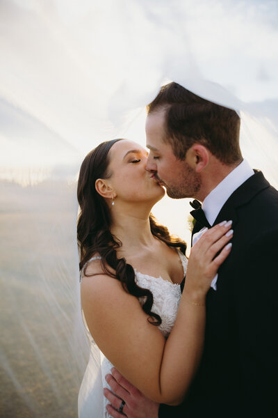 Couple shares a romantic kiss under a delicate veil, captured in a soft, intimate moment.