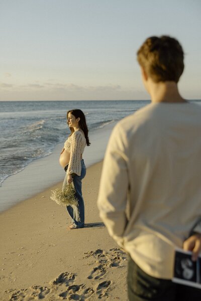 man admires his pregnant wife as she stands on the beach with flowers