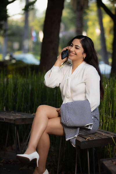 Sacramento branding photography photoshoot with woman posing on the phone sitting on a bench at the park. She is dressed in business attire and smiling as she appears to be on a business call.