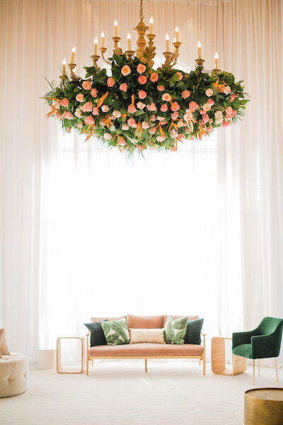 BP Collective modern lounge with blush sofa, emerald chairs, and statement chandelier adorned with peach roses and greenery.