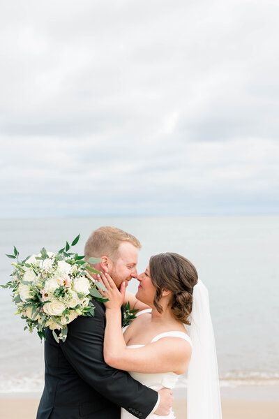 Bride and groom belly button to belly button with bouquet around grooms neck with the ocean behind them
