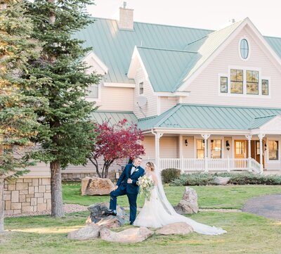 A couple stands outside of a venue in Denver while the groom puts one foot on a rock and the bride wraps her arm through his arm