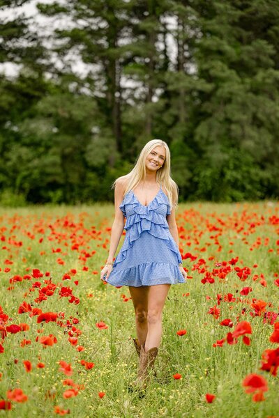 blonde charlotte area graduate walks amongst the red poppy flowers wearing a blue dress and boots