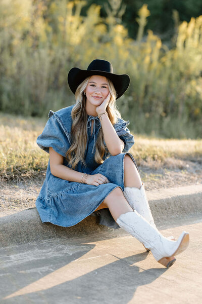 senior portrait of girl in denim dress, black cowboy hat, and white boots sitting on East Texas curb