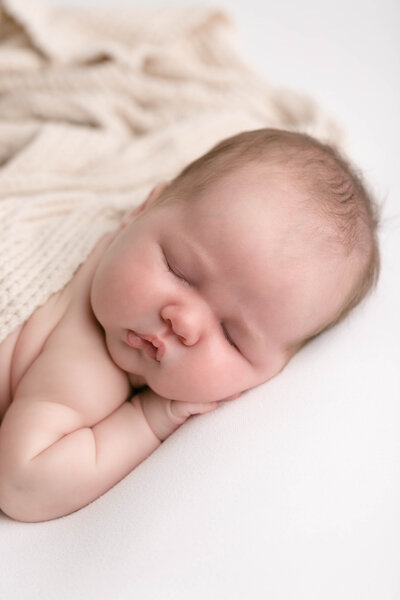 Light-skinned newborn baby sleeping on white blanket with a cream textured blanket wrapped over her. Sleeping peacefully.