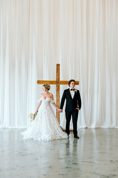 Elegant bride holding a tulip bouquet in a sleek, modern white gown, standing beside the groom in a classic black tuxedo. A serene moment from a faith-centered wedding planned by Grace-Filled Gathering Co.