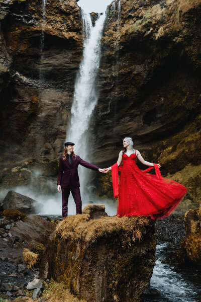 Bride in red dress and groom are standing on a mossy rock in front of a waterfall, dramatic portrait of a married couple in Iceland