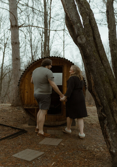 couple holding hands and walking into a rustic outdoor barrel sauna in the woods during a cozy winter elopement