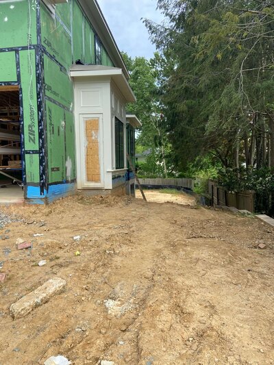 Side yard under construction with exposed dirt, an unfinished house exterior, and a retaining wall in the background.