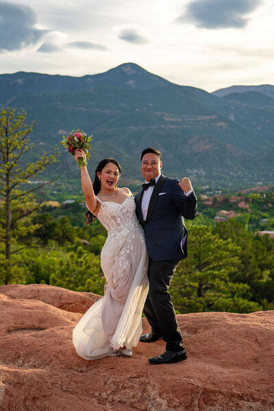 Bride holds bouquet above her head and the  groom fist bumps to the air as they celebrate their marriage at the red rocks at Garden of the Gods in Colorado Springs.