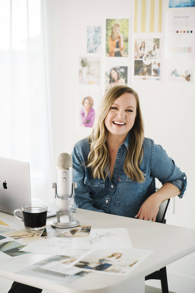 Krista Marie, a brand photographer for women, holding a cup of coffee and sitting at her desk with her podcast mic and brand photos on display