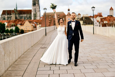 Ein gerade vermähltes Brautpaar schlendert voller Freude die Steinerne Brücke in Regensburg entlang. Die Braut streckt ihre Hand mit dem Brautstrauß weit in den Himmel.