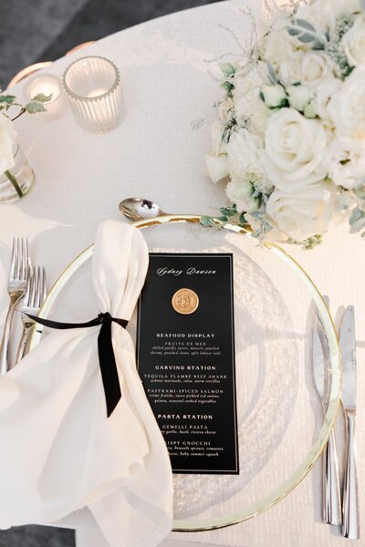 Elegant dinner table setting with a menu, white napkin, cutlery, candles, and a bouquet of white roses. This wedding was planned and designed by Thater Made, a luxury wedding and event producer based in San Diego, CA.