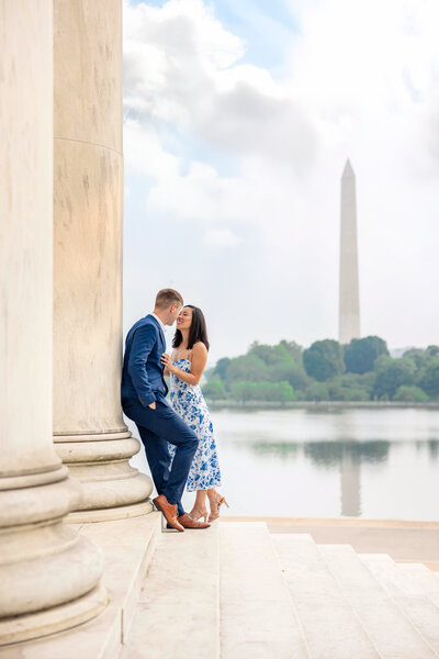 A couple shares a romantic moment by a large stone pillar, with the Washington Monument and a reflective body of water in the background. The woman wears a white and blue dress, while the man is in a blue suit. The setting appears serene and picturesque.