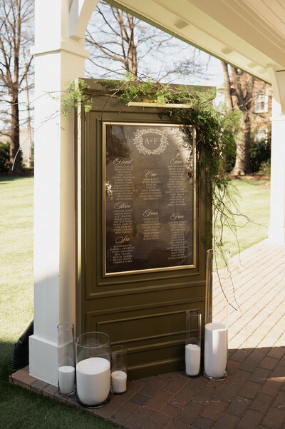 Freestanding wedding seating chart display featuring an olive green paneled structure with gold-framed mirror acrylic and white calligraphy. Topped with organic greenery and styled with pillar candles, this installation adds a refined, textural statement to an outdoor celebration.