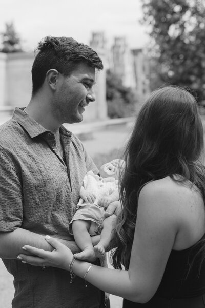Husband looks lovingly at his wife while holding their newborn baby.
