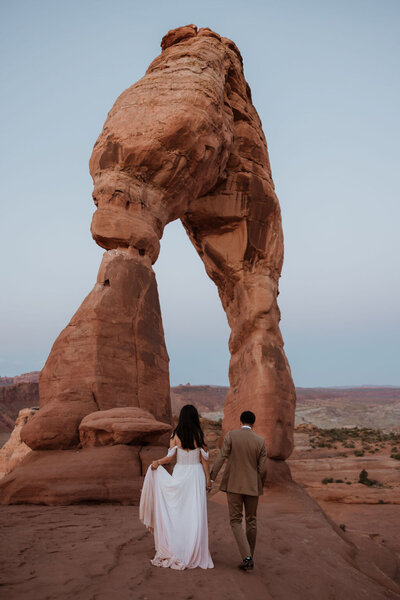 bride and groom walking towards large rock arch in the desert