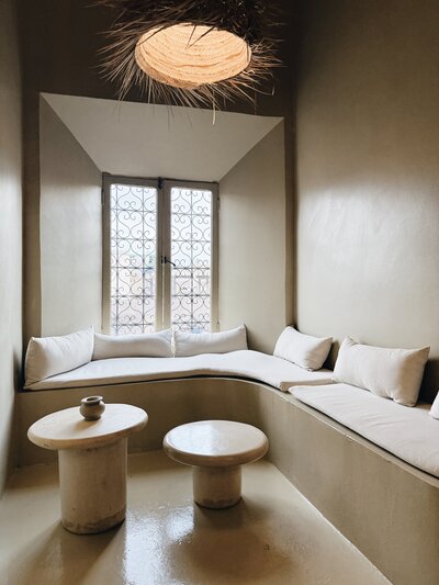 Minimalist Moroccan-inspired interior with curved built-in seating, layered white cushions, two small round stone tables, and a woven pendant light, styled in earthy neutral tones.