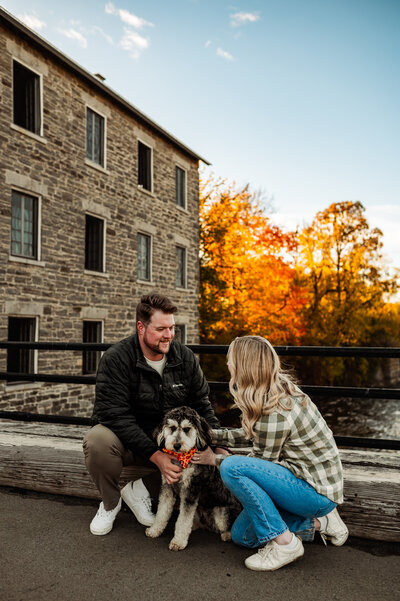 Couple with their dog sitting outside Watson’s Mill in Manotick during Ottawa fall mini session with autumn leaves in the background.