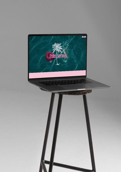  Laptop mockup displaying the Palm Springs Showit website template on screen. The homepage shows a white palm tree illustration with pink retro typography over a turquoise water background, styled with a soft pink footer. The laptop sits on a tall black stool against a minimal gray background.