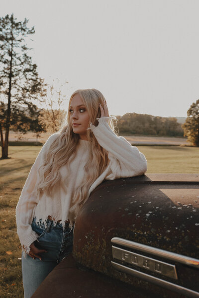 Blond senior girl standing by car in field looking away