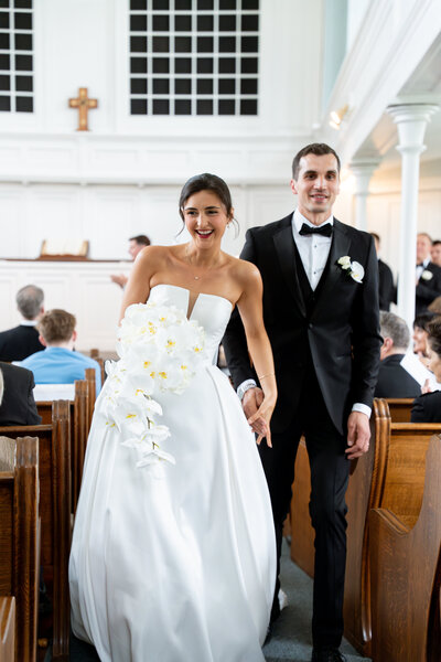 A newly married couple walks back down the aisle holding hands. The couple smiles with newly wed bliss. 