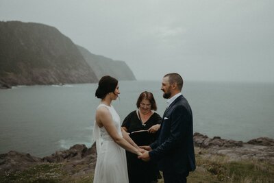 bride and groom stand on the edge of a cliff over the ocean during their Newfoundland elopement ceremony