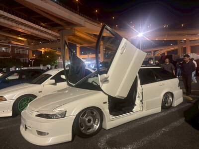 A sleek custom sports van with vertical doors open, showcased under the glowing expressway lights at the iconic Daikoku Parking Area in Tokyo.