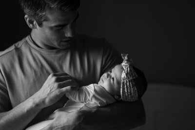 A black and white portrait captured by JPF Galleries in Fort Worth, featuring a father holding his daughter as natural light softly illuminates her face. He looks down at her with quiet love, creating a timeless, emotive moment.