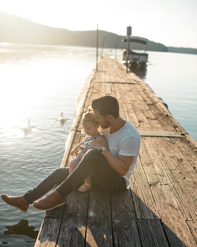 Father sitting on a wooden dock with his legs stretched out over the water, holding his child in his lap and watch the ducks swim by them in the water