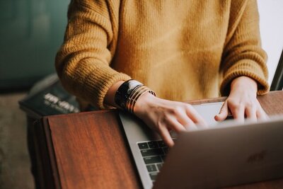 Image of a person's torso sitting at a laptop working at their computer. They are wearing a mustard colored sweater.