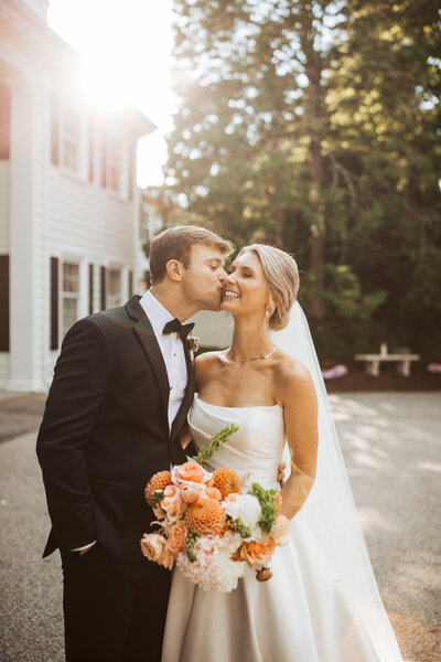 A close up of a couple with the groom kissing the brides cheek with the sun shinning behind and the wife holding her bouquet.